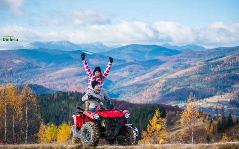 Due persone sorridenti su un quad in un paesaggio montano tranquillo, circondato da alberi dai colori vivaci.