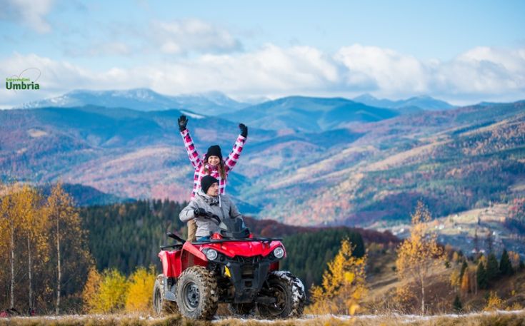 Due persone sorridenti su un quad in un paesaggio montano tranquillo, circondato da alberi dai colori vivaci.