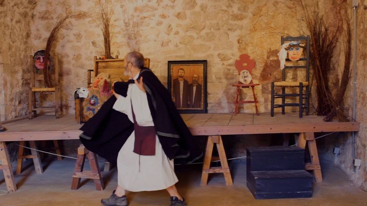 Un uomo in abiti tradizionali si muove su un palco decorato con diversi oggetti e maschere, creando un'atmosfera evocativa.