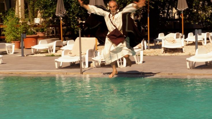 Un uomo salta in piscina indossando un abito tradizionale, mentre la luce del sole riflette sull'acqua.