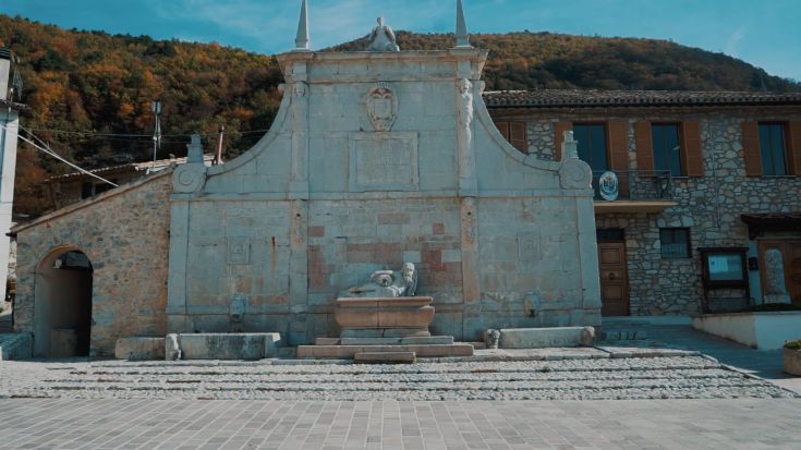 Una fontana storica, circondata da edifici in pietra e vegetazione, nel cuore di un borgo tranquillo.