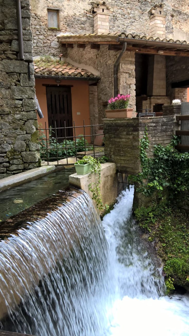 Una scena di una cascata che scorre attraverso un borgo tranquillo, circondato da vegetazione e edifici storici.