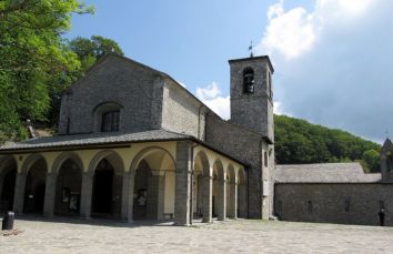 Una chiesa in pietra situata in un contesto naturale, circondata da alberi e verde intenso.