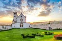 Una scena della Basilica di San Francesco ad Assisi al tramonto, con giardini verdi che la circondano.