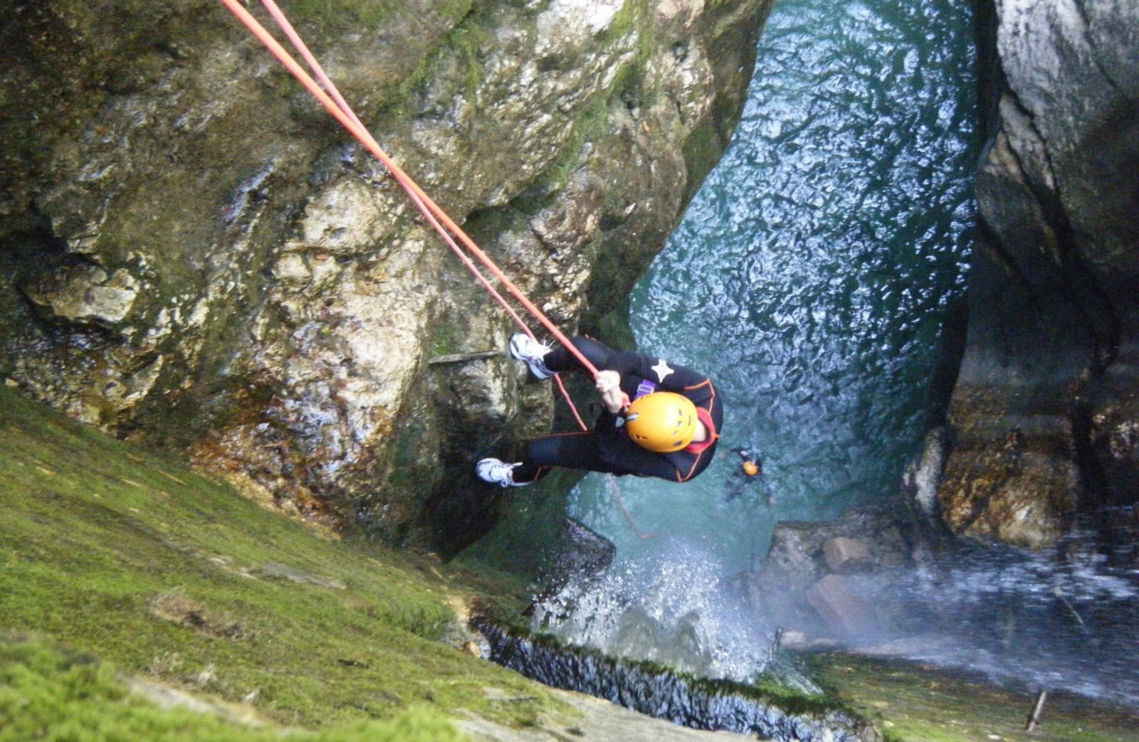  Una persona con attrezzatura sportiva e caschetto sta discendendo con una corda in una gola verso il torrente, circondato da rocce.