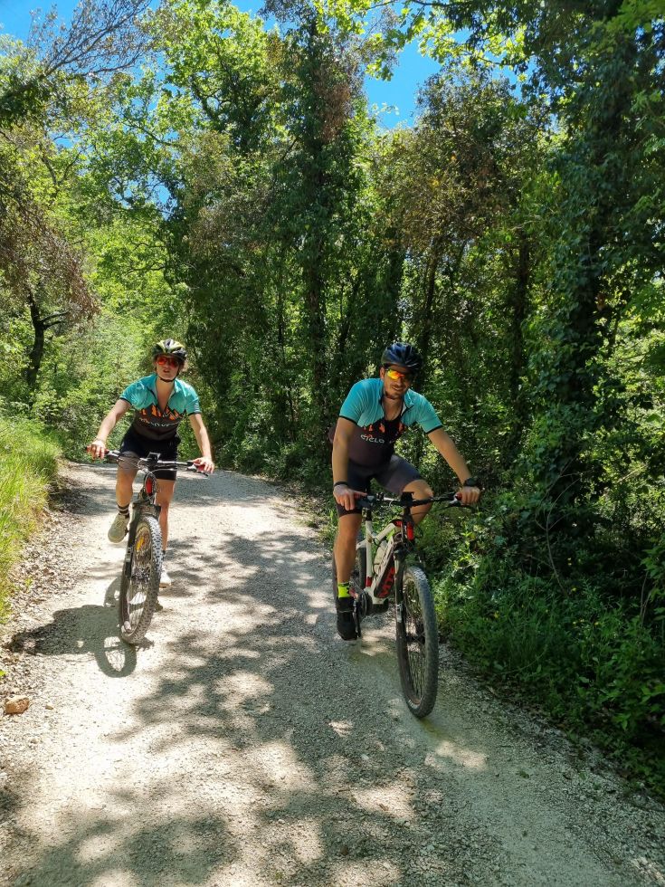 Due ciclisti percorrono un sentiero circondato da vegetazione lussureggiante e un cielo azzurro in Umbria.