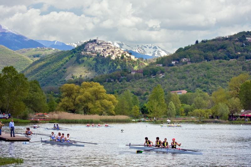 Un gruppo di persone intente in attività di canottaggio nel Lago di Piediluco, con un borgo storico sullo sfondo e montagne innevate all'orizzonte.