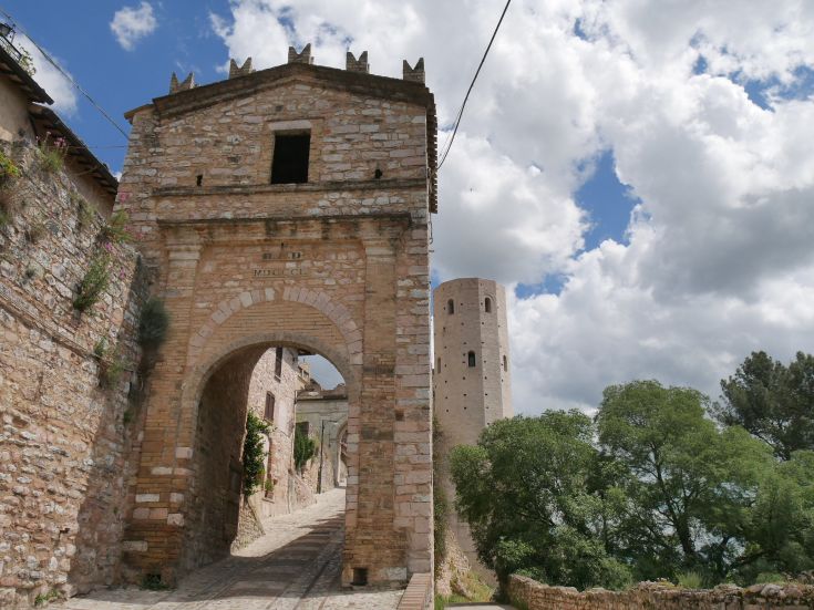 Un arco in pietra situato all'ingresso di Spello, con le torri medievali di Properzio visibili sullo sfondo.