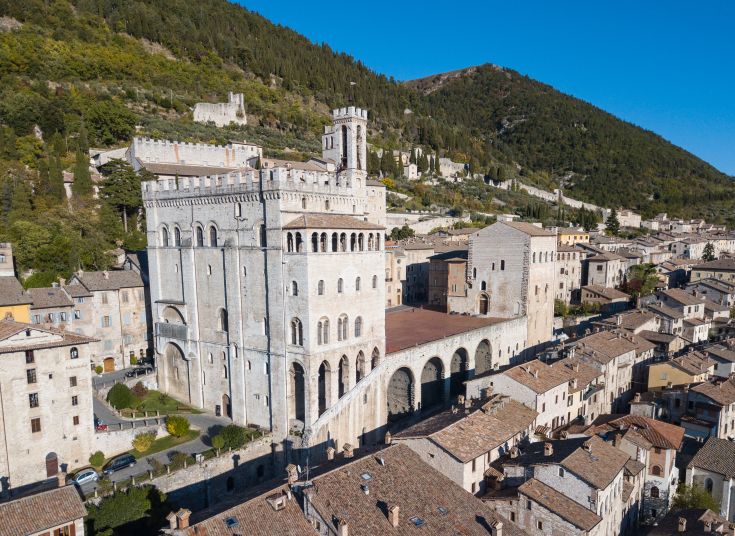Un panorama del palazzo dei Consoli di Gubbio nel centro di un antico borgo umbro, circondato da montagne e vegetazione.
