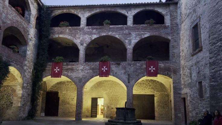 Un cortile storico caratterizzato da archi in pietra e mattoni, decorato con vasi di fiori e bandiere con il simbolo dei Cavalieri di Malta.