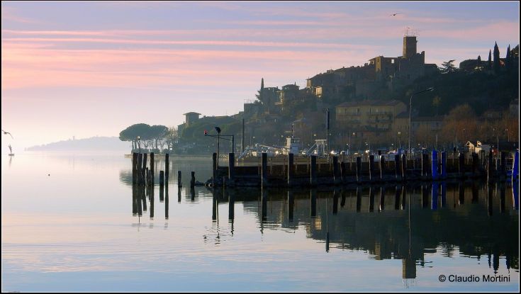 Un panorama sobrio del lago al mattino, con riflessi tranquilli e tonalità delicate nel cielo.