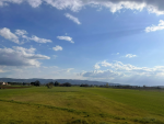 Un vasto campo verde si estende sotto un cielo sereno, punteggiato da nuvole bianche e colline in lontananza.