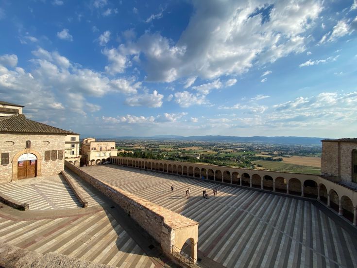 Panorama di un grande piazzale con architettura storica e un paesaggio naturale sullo sfondo.