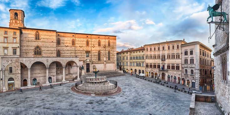 Scena di una piazza storica dell'Umbria, con eleganti edifici la fontana maggiore e cielo sereno sullo sfondo.