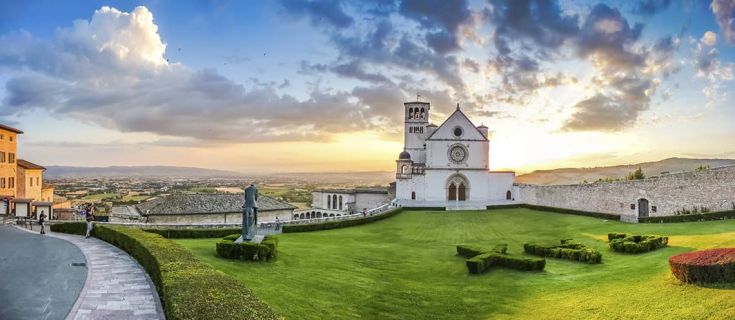 Basilica di San Francesco ad Assisi al tramonto, con il grande prato davanti alla facciata.