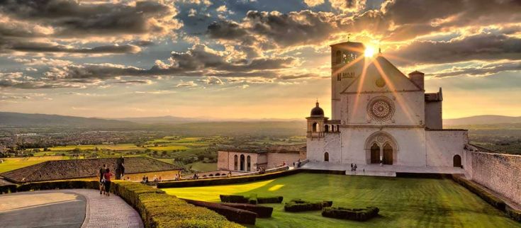 Un panorama di Assisi al tramonto, con la basilica che si staglia sotto la luce calda del sole.