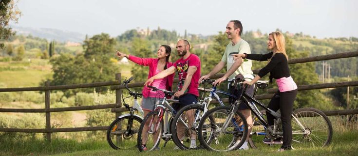 Quattro amici sorridenti in bicicletta si fermano per ammirare il panorama umbro, condividendo un momento di convivialità all'aperto.