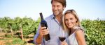 Coppia sorridente in un vigneto, con una bottiglia di vino rosso e un bicchiere in mano.
