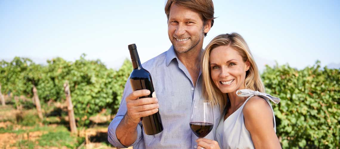 Coppia sorridente in un vigneto, con una bottiglia di vino rosso e un bicchiere in mano.