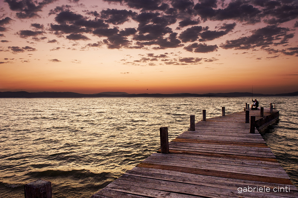 Un molo in legno si estende su un lago tranquillo al tramonto, con due pescatori che attendono di pescare.
