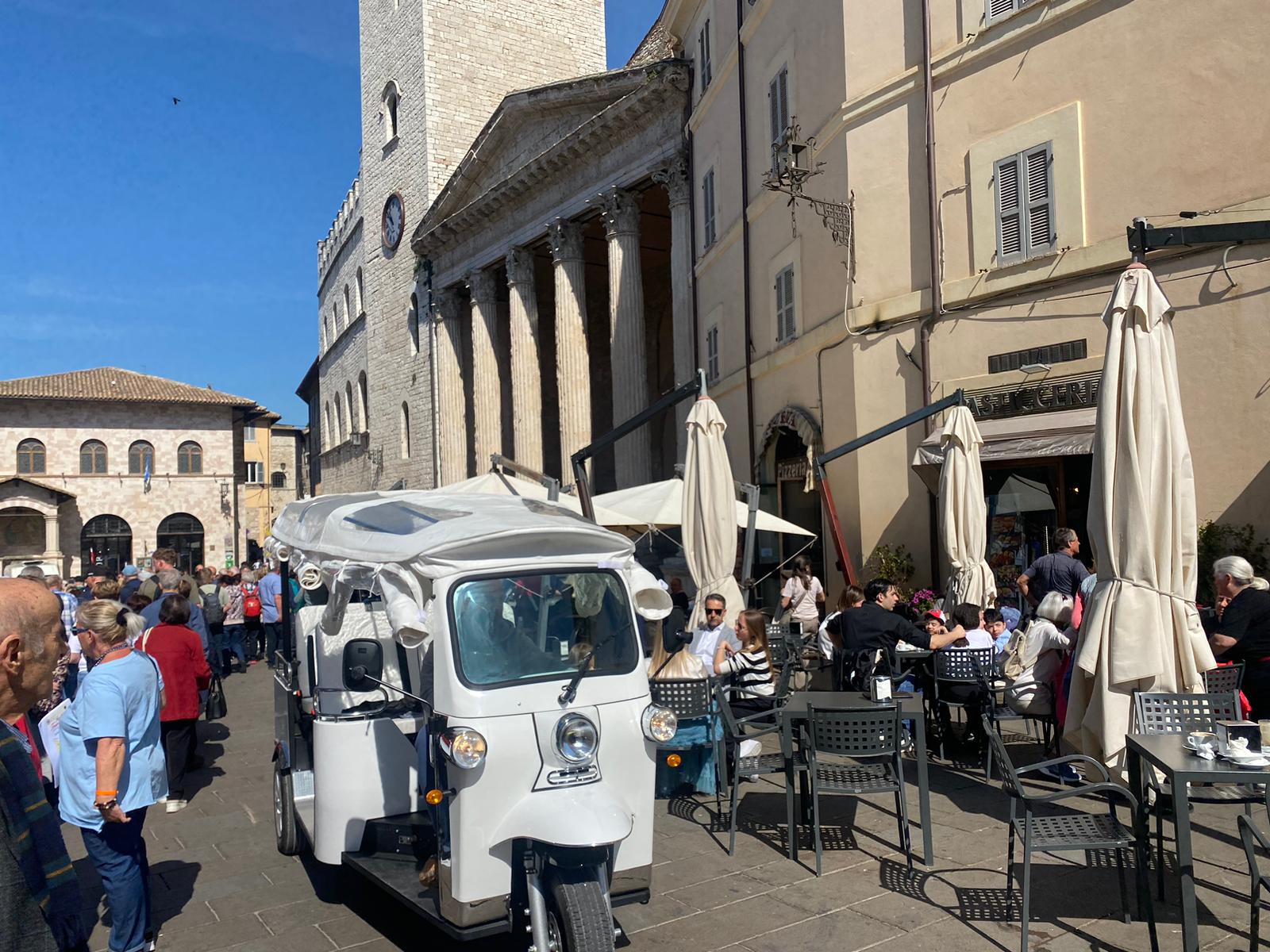 Un affollato pomeriggio di festa in Piazza di Assisi, con un'ape calessino in evidenza e visitatori che godono dell'atmosfera vivace.