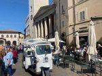 Un affollato pomeriggio di festa in Piazza di Assisi, con un'ape calessino in evidenza e visitatori che godono dell'atmosfera vivace.