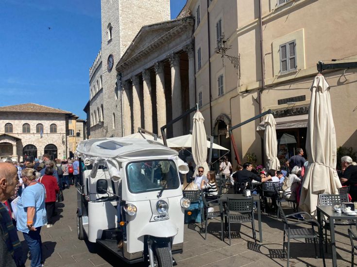 Un affollato pomeriggio di festa in Piazza di Assisi, con un'ape calessino in evidenza e visitatori che godono dell'atmosfera vivace.