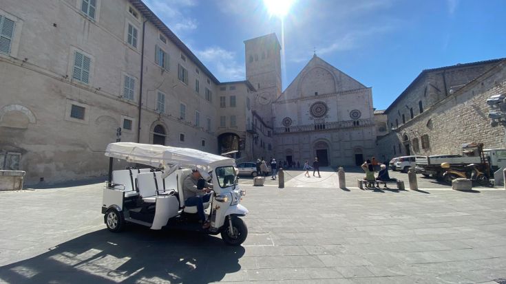 Un ape calessino bianco si ferma in una piazza, con una chiesa in stile romanico sullo sfondo.