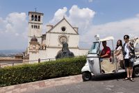 Un gruppo di persone a bordo di un ape calessino attraversa una strada di Assisi, con la basilica sullo sfondo.