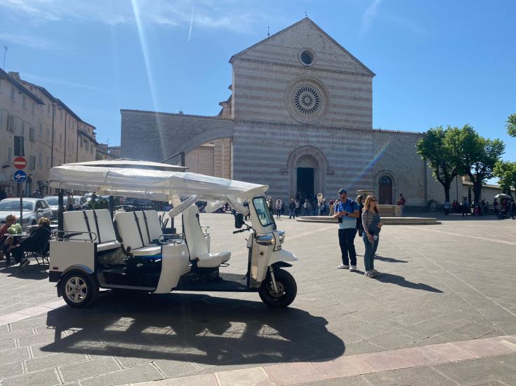 Un tuk-tuk bianco in primo piano davanti a una chiesa storica in Umbria.