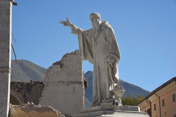 Statua in pietra di un santo che indica verso l'alto, con rovine storiche sullo sfondo.