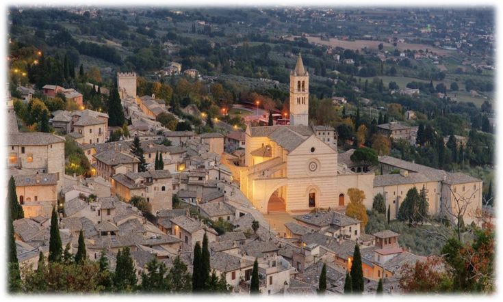 Scena di Assisi al crepuscolo, con la basilica illuminata al centro. L'atmosfera è serena e suggestiva.