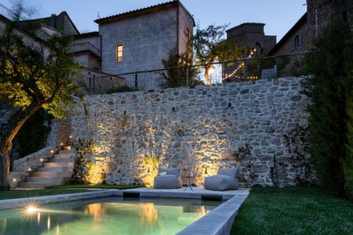 Una tranquilla area all'aperto con piscina, illuminata per la sera, circondata da un muro in pietra e piante.