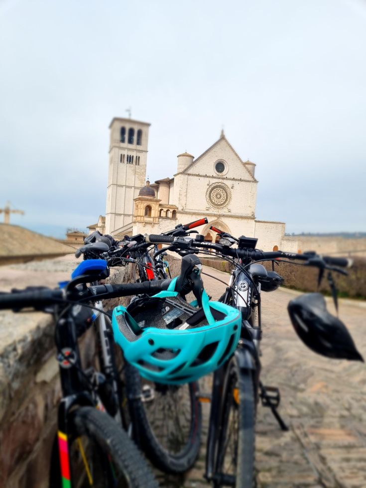 Biciclette parcheggiate davanti a un palazzo religioso, circondate dal tipico paesaggio umbro.