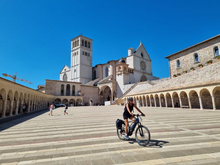 Una ciclista pedala attraverso una piazza con edifici storici in una giornata di sole.