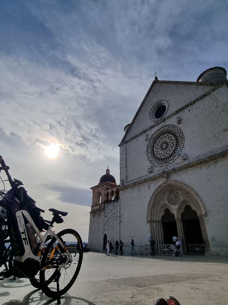 Due biciclette parcheggiate in una piazza antistante una chiesa, sotto un cielo parzialmente nuvoloso.