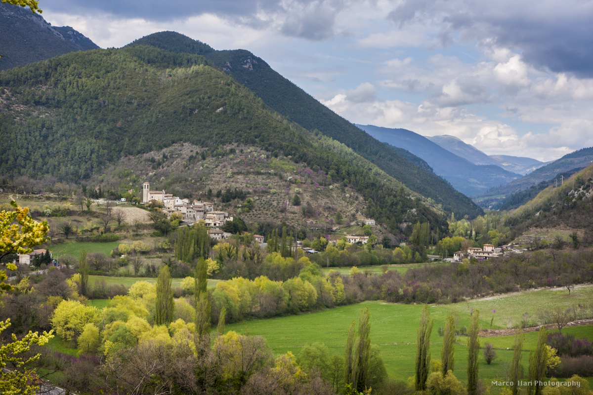 Panorama di un paesaggio umbro con un borgo storico. Montagne verdi e un cielo con nuvole caratterizzano l'ambiente.
